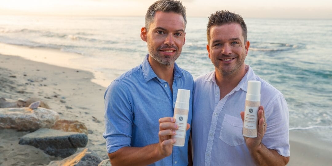 Two men at the beach holding bottles of Neora's Sunless Tanner in front of them.