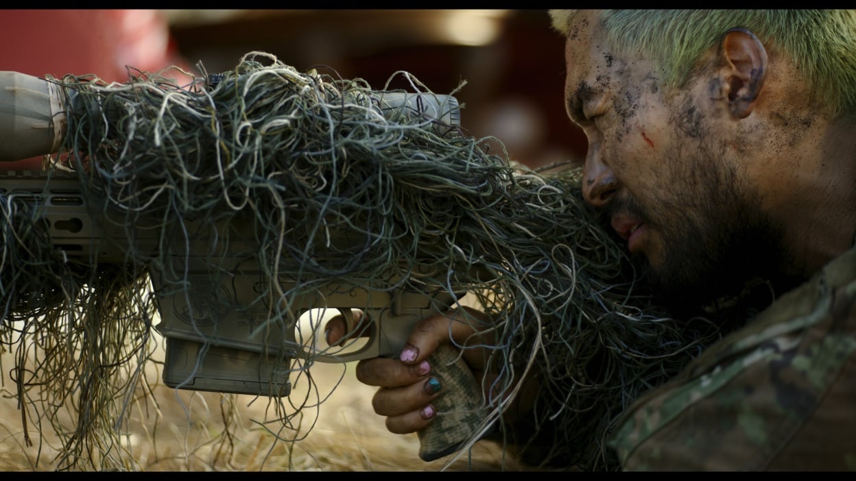 A camouflaged man aiming a sniper rifle.