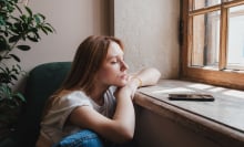 Upset redhead teen girl sitting by window looking at phone waiting call from boyfriend, feeling sad and depressed teenager looking at smartphone wait for message