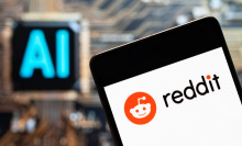 A phone displaying the Reddit logo in front of a screen that reads "AI". 