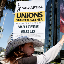 A woman stands on a sunny street holding a picket sign, while a Netflix billboard is visible in the background.