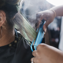 A close up of a black hairdresser straightening a client's hair with some steam rising from the heat.