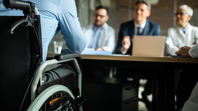  A person in a wheelchair sits across a desk interviewing with three people for a job.