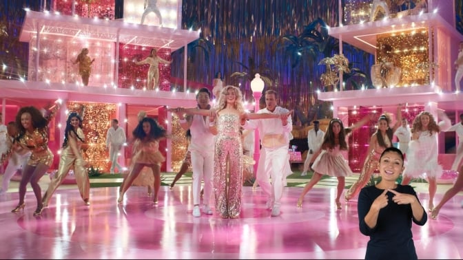 A group of coordinated Barbies and Kens dance on a brightly lit, pink dance floor. A woman in a black shirt interprets in ASL in the bottom right corner of the screen.