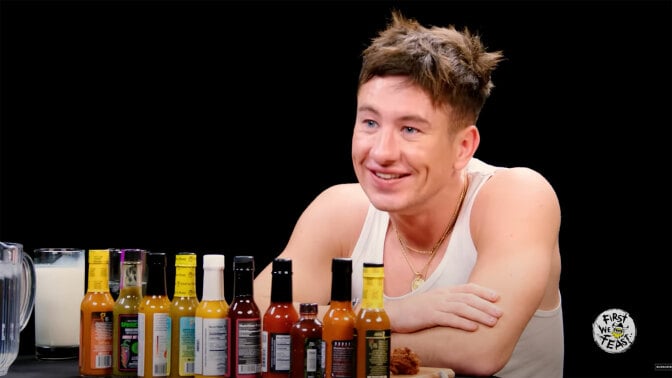 A man in a white vest sits at a table covered in sauce bottles in a dark room.