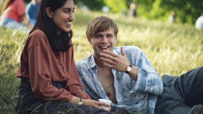 Dexter (Leo Woodall) and Emma (Ambika Mod) are both lying on grass in a park, both laughing. 