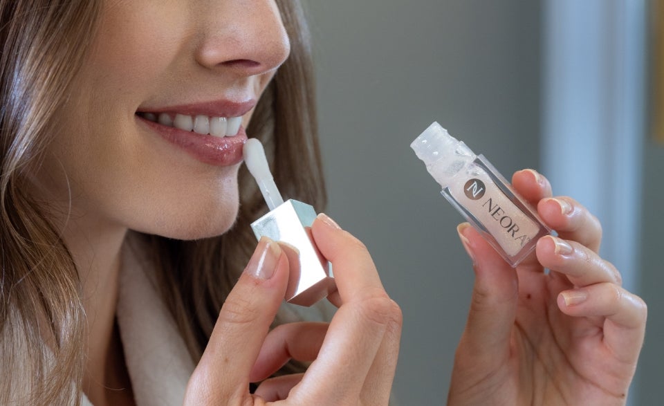 A close-up of a smiling woman applying Neora’s Shimmer Lip Oil to her bottom lip.