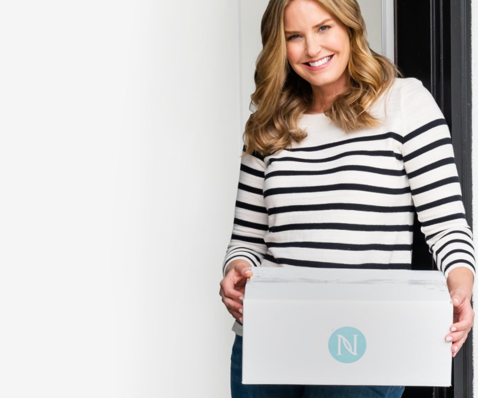 Image of woman in her front doorway smiling as she holds a white Neora box with blue logo that has been delivered.