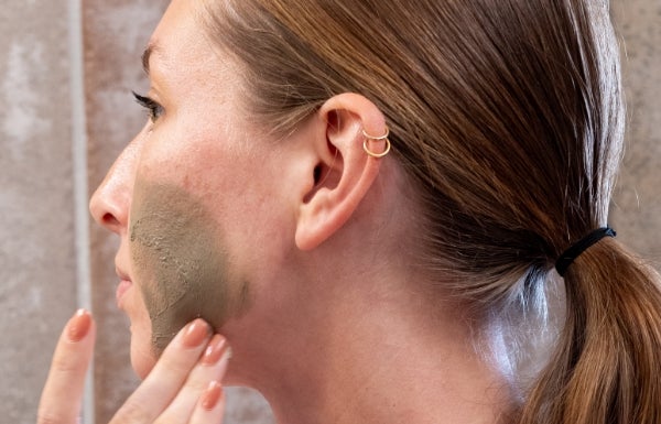 A close up of a pony-tailed woman applying Neora’s Clarifying Clay Mask on her left cheek.