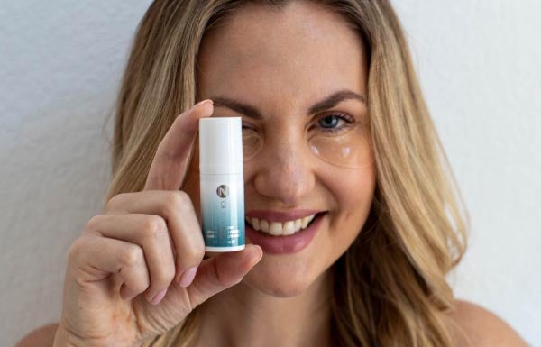 A blonde woman holding up an Eye Serum bottle while wearing Eye Patches under her eyes.