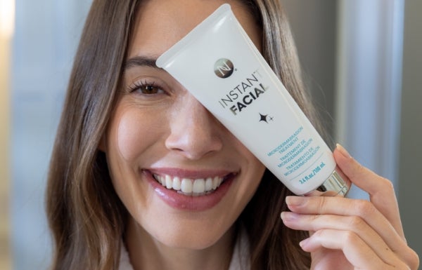 A close-up of a smiling woman holding a tube of Instant Facial over her left eye.