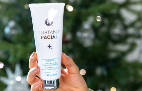 A close-up of someone’s hand holding a tube of Instant Facial in front of a festive tree.