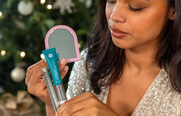 A woman holds up a tube of LashLush, Lip Pumping Serum and a pink Purse Mirror in front of a festive tree.