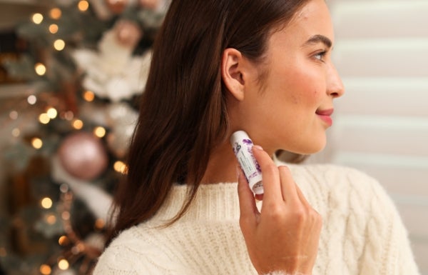 A side view of a woman applying Neora’s Zen + Calm Lavender Balm on her neck. Behind her is a festive tree.