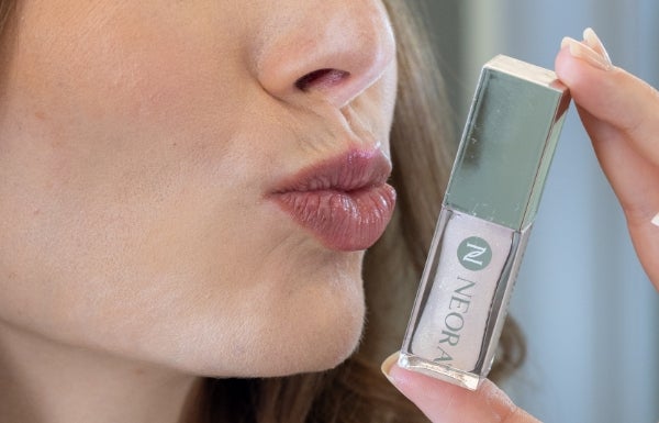 A close-up of a woman pouting next to a container of Neora’s Shimmer Lip Oil.