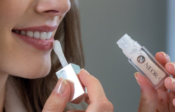 A close-up of a smiling woman applying Neora’s Shimmer Lip Oil to her bottom lip.