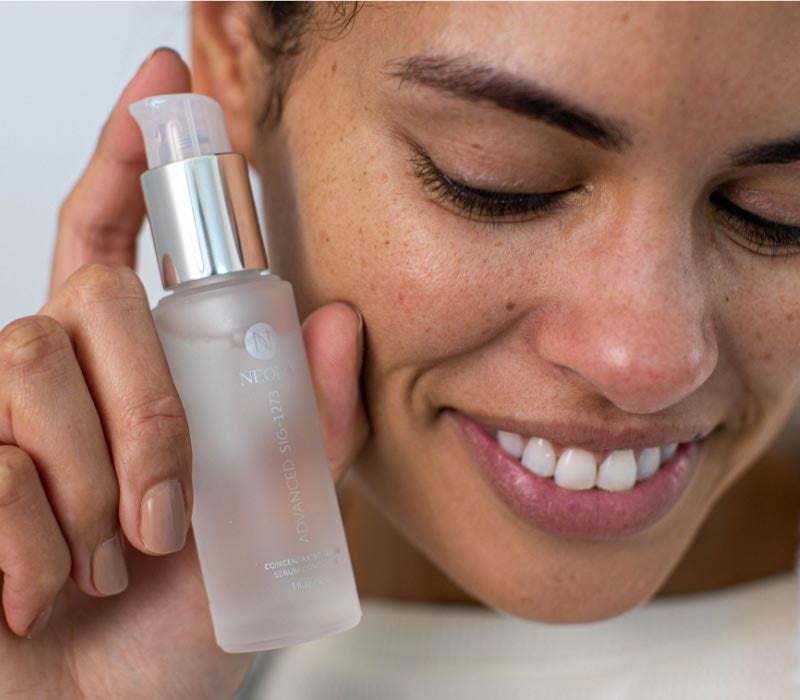 Woman holding Neora’s Advanced SIG-1273 Concentrated Serum 
                    in her hand next to her face while smiling 