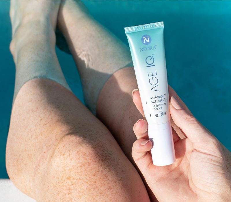 Mujer sosteniendo Neora Age IQ® Invisi-Bloc Protector Solar Gel SPF 40 en sus manos mientras está sentada en una piscina