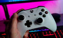 a close-up of a person holding an xbox controller in front of.an RGB keyboard and a gaming monitor