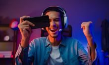 Happy asia man gamer wear headphone competition play video game online with smartphone colorful neon light in living room at night modern house. Esport streaming game online, Home quarantine activity.