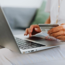 a close-up of a woman in a white top, white pants, and gold jewelry holding a credit card in front of an open macbook